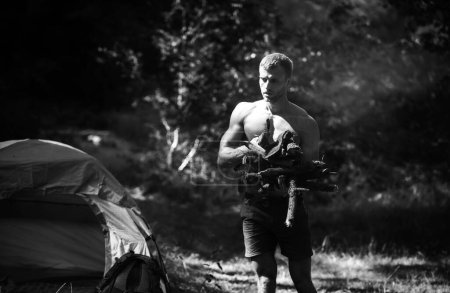 Foto de Fuerte hombre sexy con leña para hoguera. Camping al aire libre en la naturaleza. Hombre leñador guapo y musculoso con un hacha, acampando. Caminata de fin de semana - Imagen libre de derechos