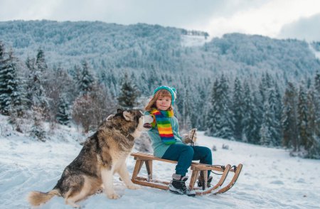 Foto de Niño con perro disfrutando de un paseo en trineo. Niño en trineo. El niño juega afuera en la nieve. Invierno, vacaciones y Navidad tema - Imagen libre de derechos