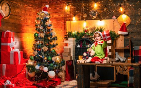 Foto de Happy little boy está jugando con juguetes junto al árbol de Navidad y mostrando su regalo de Navidad a la cámara. El niño lleva ropa de Santa Claus. Concepto de vacaciones - Imagen libre de derechos