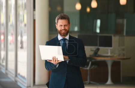 Téléchargez les photos : Homme d'affaires tenant son ordinateur portable debout en costume sur fond de bureau - en image libre de droit