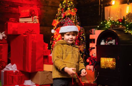 Foto de Feliz niño de Navidad con sombrero de Santa en el fondo del árbol de Navidad. Concepto de vacaciones. Niño con regalo de Navidad en sus manos - Imagen libre de derechos