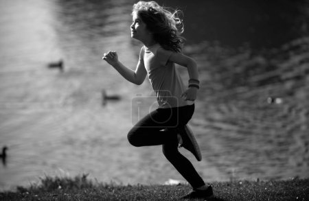 Téléchargez les photos : Enfant garçon courir ou faire du jogging près du lac sur l'herbe dans le parc. Petits coureurs faisant du jogging en plein air dans la nature estivale. Enfants courant sur la prairie verte contre la mer ou le lac - en image libre de droit