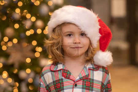 Photo for Close up portrait of child in Santa hat near Christmas tree at home. Cute Child portrait near Christmas present. Kid funny face with Christmas tree. Little boy playing under Christmas tree. Winter - Royalty Free Image