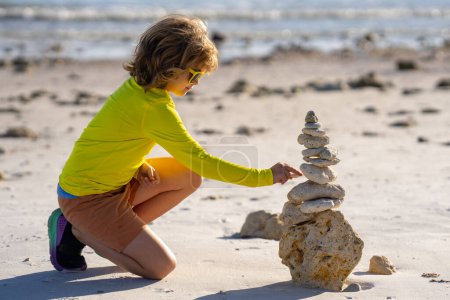 Téléchargez les photos : Calme jeu d'enfant avec des pierres pyramidales équilibre sur le sable de la plage d'été. Les enfants jouent avec une pile de pierres. Les enfants jouent sur la plage. Concept d'équilibre et d'harmonie des enfants. Jeu d'enfant en vacances d'été - en image libre de droit