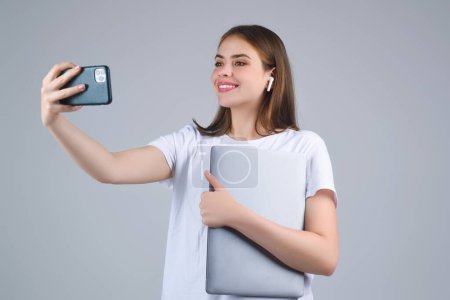 Foto de Mujer joven Estudiante en camiseta blanca sosteniendo teléfono y portátil, aislado sobre fondo gris. Estudiante haciendo videollamada. Estudiante con teléfono inteligente en el estudio. Retrato del estudiante que tiene videollamada - Imagen libre de derechos