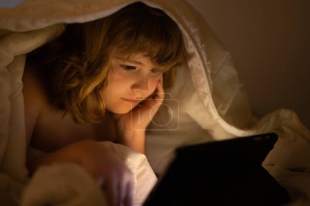 Téléchargez les photos : Enfant au lit sous une couverture joue sur une tablette dans un jeu dans le noir. Le visage de l'enfant est éclairé par une tablette. Enfant utilisant une tablette au lit à la maison. Enfant dans la chambre à coucher regarder un film ou jouer au jeu sur - en image libre de droit