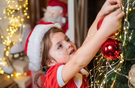 Photo for New year and Christmas kids concept. Christmas kid decorating Christmas tree with bauble. Little girl in santa hat decorating Christmas tree with baubles - Royalty Free Image