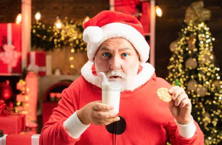 Foto de Feliz Santa Claus - con un vaso de leche y galletas. Galletas de Navidad y leche. Santa diversión. Retrato de Papá Noel sorprendido y divertido. Santa hacer cara divertida y recoger galletas - Imagen libre de derechos