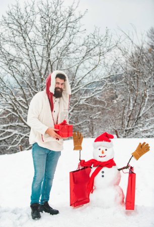 Téléchargez les photos : Bonhomme de neige souriant heureux par une journée d'hiver ensoleillée avec père heureux. Mignon petit bonhomme de neige et homme barbu avec sac à provisions. Bonhomme de neige drôle avec sac à provisions - rabais et concept de vente d'hiver - en image libre de droit