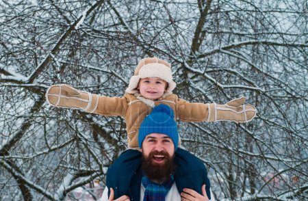 Foto de El niño se sienta sobre los hombros de su padre. Padre llevando a su hijo de vuelta al parque. Feliz Navidad y Feliz Año Nuevo. La mañana antes de Navidad - Imagen libre de derechos