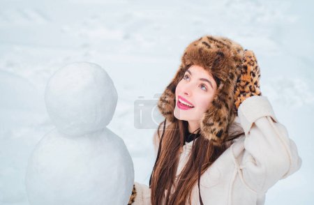 Foto de Joven bella alegre divirtiéndose con muñeco de nieve en Winter Park. Divertidos muñecos de nieve. Feliz chica sonriente hacer muñeco de nieve en el día de invierno soleado - Imagen libre de derechos