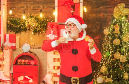 Téléchargez les photos : Portrait du Père Noël Boire du lait dans du verre et tenir des biscuits. Style barbe de Noël. Joyeux Noël. Père Noël au chapeau de Père Noël. Nourriture et boisson de Noël - en image libre de droit
