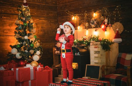 Photo for Happy little kid is wearing Santa clothes, with Christmas gift box having an idea. Christmas concept. Fireplace background. Holidays - Royalty Free Image
