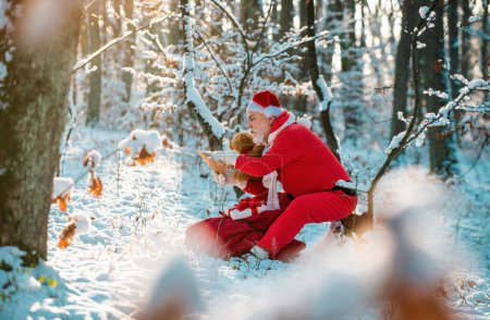 Téléchargez les photos : Noël Santa lettre de lecture près de l'arrière-plan de la nature de Noël. Funny Santa in Santa chapeau lire lettre sur fond de nature d'hiver - en image libre de droit