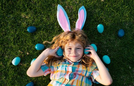 Kid boy hunt egg on Easter day outdoor. Top view funny kids laying on grass. Kids Easter bunny hunting eggs, wear rabbit ears. Spring family holiday