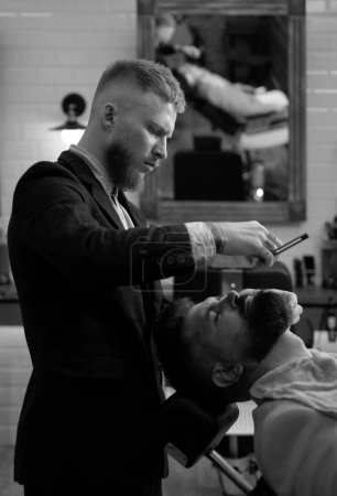 Téléchargez les photos : Homme barbu visitant coiffeur dans le salon de coiffure. Un barbu se fait couper les cheveux par un coiffeur au salon de coiffure. Coiffeur et coiffeur - en image libre de droit