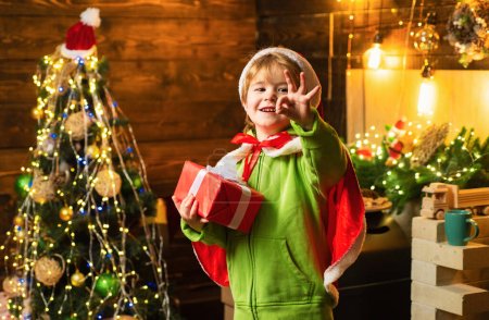 Foto de Niño feliz junto al árbol de Navidad mirando a la cámara con regalo de Navidad en sus manos. Niño pequeño lleva ropa de Santa Claus - Imagen libre de derechos