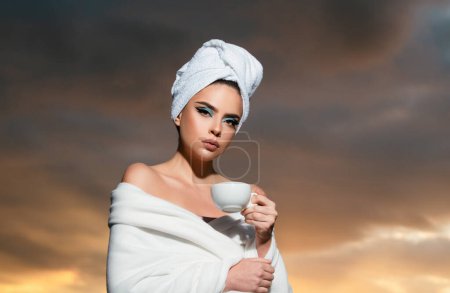 Téléchargez les photos : Portrait of woman in bathrobe holding a cup with coffee at sunrise. Beauty woman with towel on head drinking coffee in the sun, outdoor in sunlight light, enjoying morning coffee - en image libre de droit