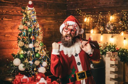 Téléchargez les photos : Joyeux Père Noël habillé en vêtements d'hiver pensez à Noël près de l'arbre de Noël. Portrait d'un Père Noël mature brutal - en image libre de droit