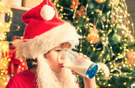 Téléchargez les photos : Joyeux Père Noël qui mange un cookie et boit un verre de lait à la maison. Joyeux Noël. Enfant Père Noël profiter dans le gâteau au pain d'épice servi et le lait - en image libre de droit