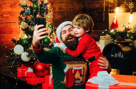 Téléchargez les photos : Heureux père et fils parlant avec téléphone portable près de l'arbre de Noël. Petit Père Noël et vieux Père Noël barbu en utilisant un smartphone. Internet, sans fil et concept de médias sociaux - en image libre de droit
