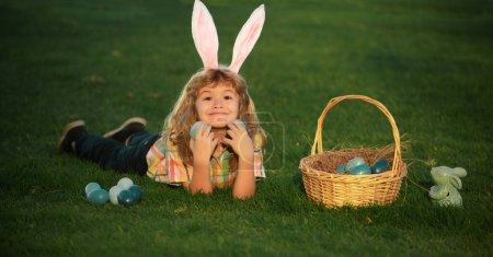 Téléchargez les photos : Child boy hunting easter eggs in spring lawn laying on grass. Bunny kids with rabbit bunny ears - en image libre de droit
