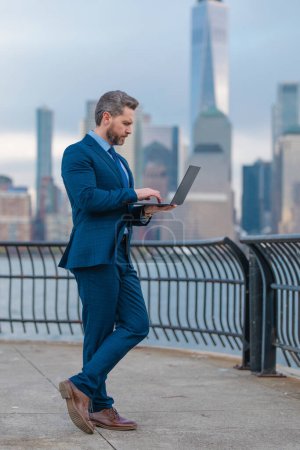 Photo for Businessman in suit holding laptop on urban street in NYC. Successful business man outdoor. Middle aged business man standing outside office using laptop. Street portrait of mature businessman - Royalty Free Image