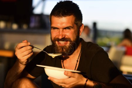Photo for Man eat soup. Good Appetite. Bearded man with bowl of soup. Happy guy eating soup outdoor. Man eating delicious soup in restaurant. Dinner and meal. Man eating lunch in a cafe - Royalty Free Image