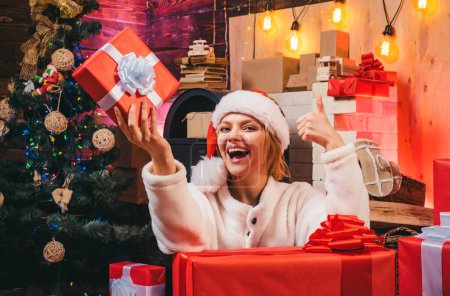 Foto de Mujer sonriente decorando el árbol de Navidad en casa. Abre la boca. Feliz Navidad. Mujer feliz. Decoraciones navideñas y caja de regalo sobre fondo de madera. Vestidos de Navidad - Imagen libre de derechos