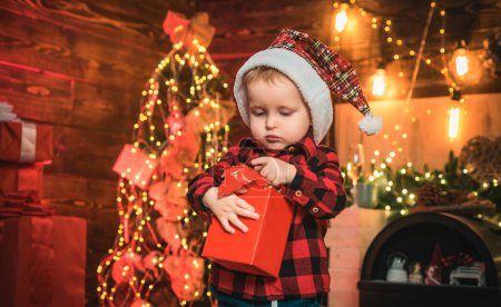 Téléchargez les photos : Petit bébé avec cadeau de Noël dans les mains sur le fond de Noël. Arbre de Noël. Heureux gamin - en image libre de droit