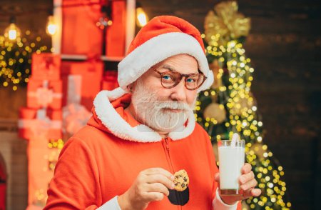 Foto de Retrato de un hombre barbudo y divertido disfrazado de Santa. Santa diversión. Papá Noel comiendo galletas y bebiendo leche en Nochebuena. Barba de Navidad estilo - Imagen libre de derechos