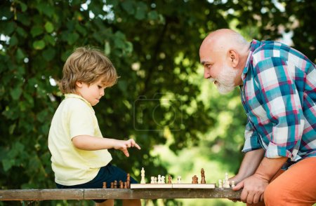Téléchargez les photos : Beau grand-père et petit-fils jouent aux échecs tout en passant du temps ensemble. Petit garçon jouant aux échecs avec son grand-père - en image libre de droit