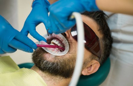 Photo for Handsome man at dentists office. Female dentist checking patient teeth with mirror in modern dental clinic. Examine of young man by dentist on light blurred background - Royalty Free Image