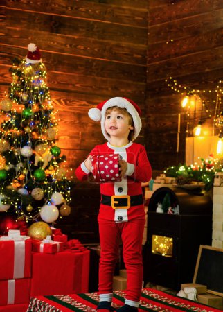 Foto de Feliz niño decorando el árbol de Navidad. Lindo niño pequeño cerca del árbol de Navidad. Decoraciones navideñas. Los niños disfrutan de las vacaciones. Niño de Navidad sosteniendo una caja de regalo roja - Imagen libre de derechos