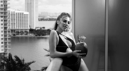 Téléchargez les photos : Sexy jeune femme blonde en maillot de bain avec cocktail de noix de coco posant sur le balcon vue sur la ville. Vacances d'été à Miami. Boisson à la noix de coco. Vacances tropicales - en image libre de droit