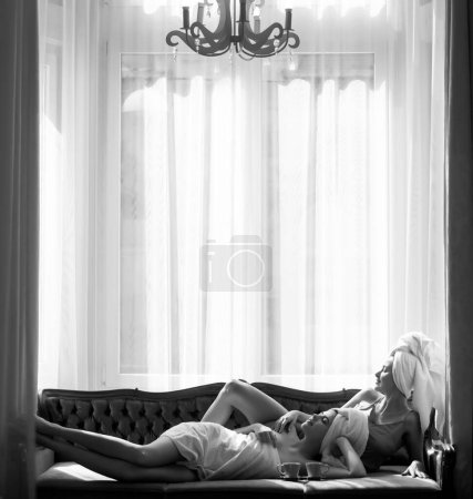 Foto de Dos mujeres hermosas jóvenes en batas blancas y toallas en la cabeza en la cama después de despertar. Chicas fritas en la cama en lujoso apartamento rico o habitación de hotel. Descanso matutino - Imagen libre de derechos