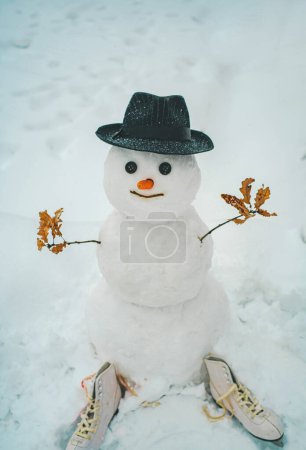 Téléchargez les photos : Bonhomme de neige drôle avec une carotte au lieu d'un nez et dans un chapeau tricoté chaud sur une prairie enneigée sur un fond de neige floue. Bonhomme de neige dans un foulard et un chapeau. - en image libre de droit