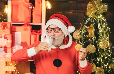 Foto de Papá Noel comiendo galletas y bebiendo leche. Santa Claus sosteniendo galletas de Navidad y leche sobre el fondo del árbol de Navidad. Galletas de Navidad y leche. Saludo tarjeta de Navidad - Imagen libre de derechos