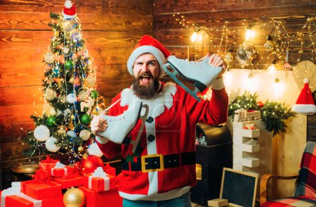 Téléchargez les photos : Portrait de mode d'un bel homme à l'intérieur avec arbre de Noël. Nouvel an concept de Noël. Mauvais Père Noël au chapeau de Père Noël. Joyeux Noël et bonne année - en image libre de droit
