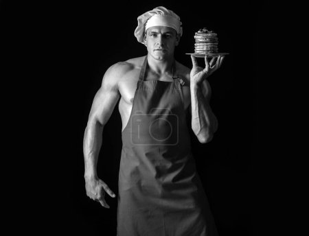 Photo for Sexy man in chefs hat with sweet homemade stack of pancakes with syrup isolated on black. Chef muscle man with apron bake pancakes with berries and maple syrup - Royalty Free Image