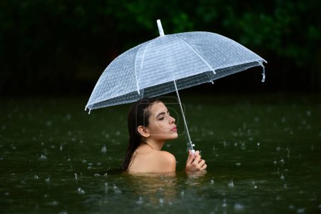 Téléchargez les photos : Jeune femme sexy dans le lac pluvieux ou l'eau de mer tenir parapluie avec gouttes d'eau de pluie. Fille sous les gouttes d'eau tombant en saison des pluies. Femme sensuelle avec parapluie. Pluie estivale. Météo pluvieuse - en image libre de droit