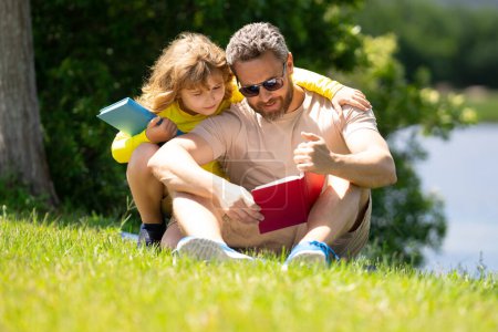 Photo for Father and son sitting on green grass in garden and reading book together. Happy family reading book together in green nature. Father is sitting with son read book. Kid is very interested - Royalty Free Image