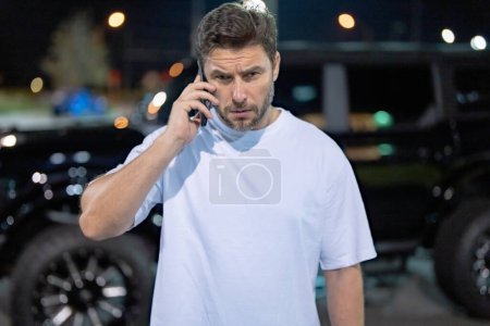 Photo for Angry man talking on phone near car on night urban street. Dangerous aggressive man talking on phone with serious face. Criminal city. Danger district. Aggressive angry man talking on phone outdoor - Royalty Free Image