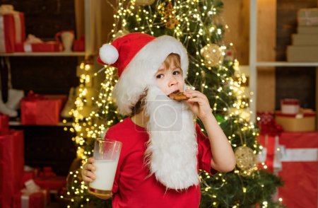 Téléchargez les photos : Joyeux Père Noël - petit garçon avec un verre de lait et des biscuits. Portrait de Père Noël surpris et drôle. Biscuits pour enfants Père Noël. Père Noël. Enfant de Noël - en image libre de droit