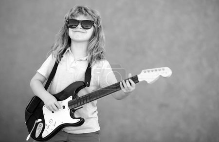 Foto de Funny rock child with guitar. Little boy in sunglasses. Child musician guitarist playing electric guitar. Kids music concept - Imagen libre de derechos
