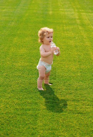Téléchargez les photos : Bébé boire de l'eau propre de la bouteille en plein air sur le champ vert printemps. Enfant en bonne santé - en image libre de droit