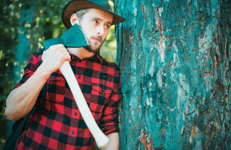 Foto de Woodcutter with axe in the summer forest. Lumberjack standing with axe on forest background. Lumberjack in the woods with an ax - Imagen libre de derechos