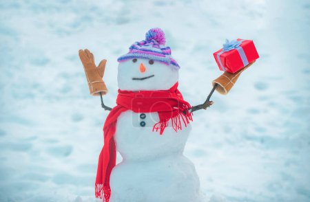 Foto de Lindo muñeco de nieve en sombrero y bufanda en el campo cubierto de nieve con regalo de Navidad sorpresa. Muñeco de nieve con bolsa de compras - concepto de compra de regalos. Feliz hombre de nieve sonriente - venta concepto de descuento - Imagen libre de derechos
