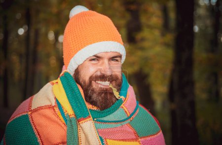 Téléchargez les photos : Joyeux hipster homme à la barbe enveloppée dans une couverture multicolore, coiffé d'un chapeau chaud, souriant à la caméra. Concept de résistance aux maladies saisonnières, rhumes, prévention nez qui coule - en image libre de droit