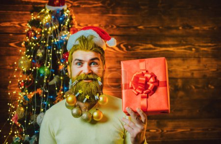 Téléchargez les photos : Portrait du beau Père Noël à l'intérieur avec cadeau de Noël. Homme barbu s'amuser près de l'arbre de Noël à l'intérieur. Père Noël à la maison avec sac de Noël - en image libre de droit